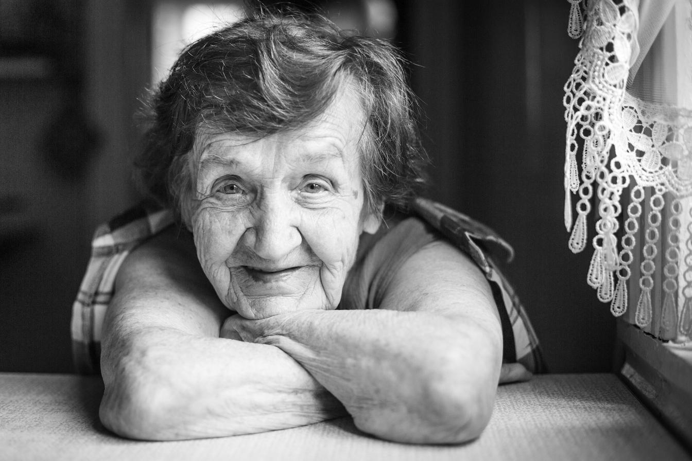 An elderly woman smiling at home, resting on her arms beside lace curtains